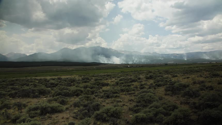 Smoke billows from a wildfire burning in a vast green valley in front of a majestic mountain range