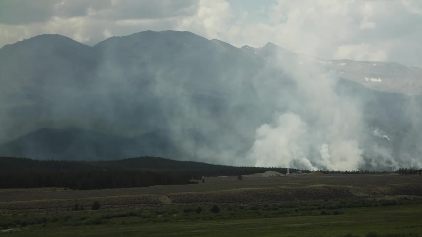 Thick white smoke rises from a wildfire burning in a vast mountain landscape