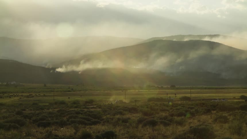 A lone car travels through a vast, hazy valley as golden sun rays illuminate the smoke from a wildfire on the distant hills