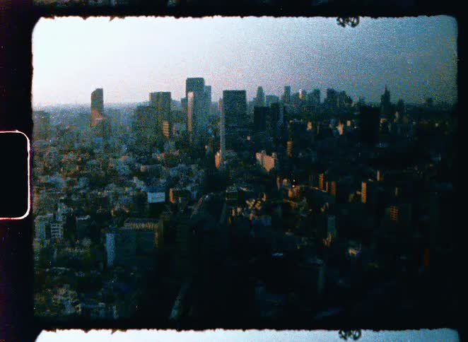 A vintage, grainy 8mm film captures a sprawling city skyline as the light of day fades