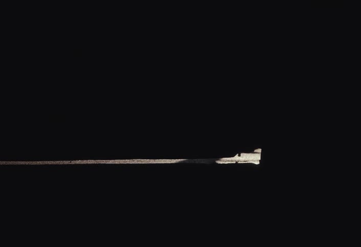 A person closes a heavy rustic door from the outside, plunging the room into darkness with only a sliver of light remaining