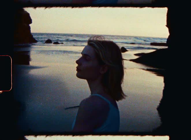 A nostalgic film-style portrait of a young woman looking out at the ocean on a serene beach during a golden sunset