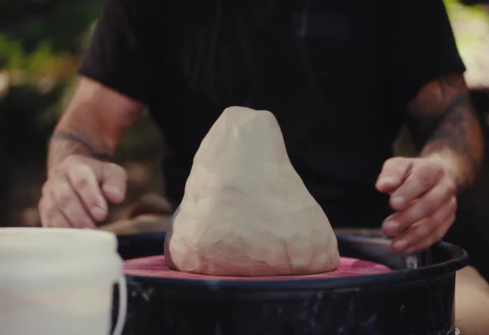 A potter with tattooed arms slams a lump of fresh clay onto a pottery wheel to begin shaping it