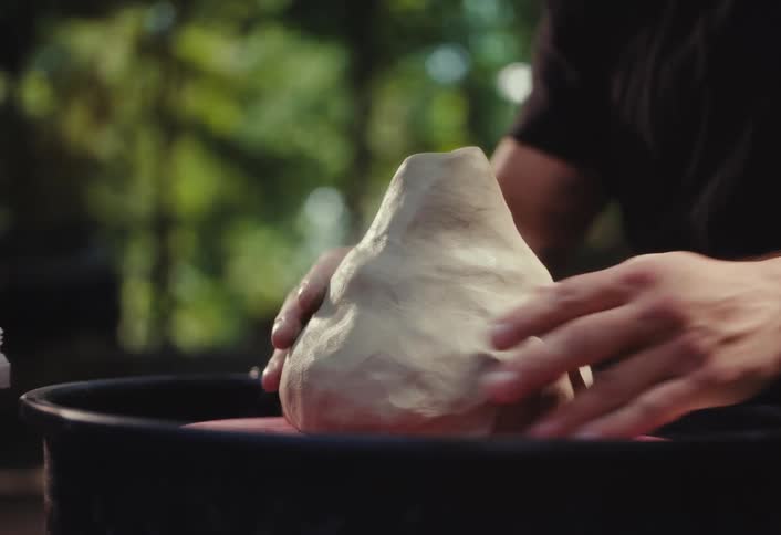 A potter's hands knead and place a lump of raw clay onto a potter's wheel outdoors