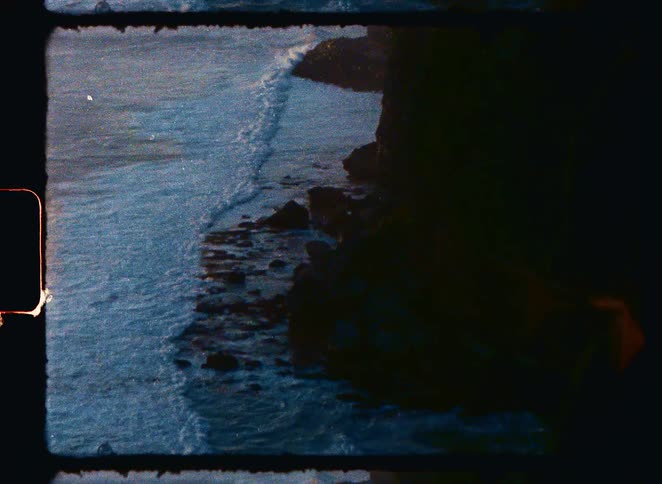 Authentic vintage 8mm footage shows a tranquil scene of ocean waves rolling onto a rocky coastline
