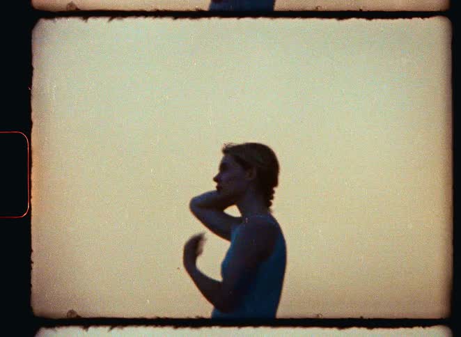 A silhouetted woman on a beach ties her hair back and gazes at the ocean waves during a warm sunset, captured on vintage 8mm film