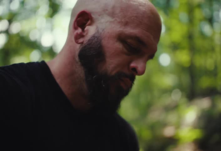 A focused, tattooed artisan with a beard kneads a lump of clay in a sunlit outdoor workshop