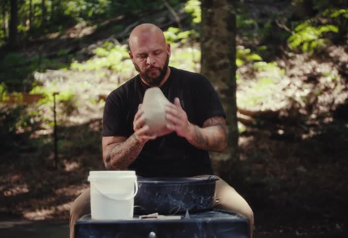 A bald, bearded potter prepares to throw clay on a wheel set up in a peaceful, sunlit forest