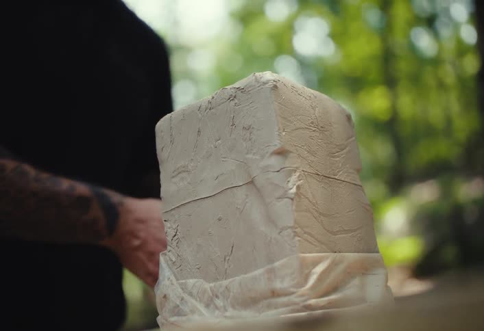 A sculptor with tattooed hands carefully cuts a large block of wet clay with a wire tool in his outdoor workshop