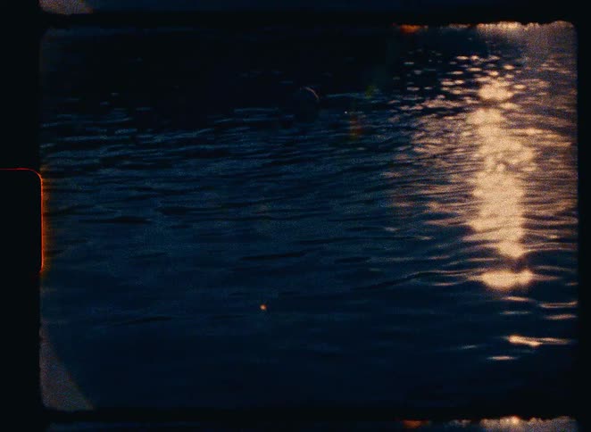 Vintage film captures the golden reflection of a sunset shimmering on dark, rippling water where a person is swimming
