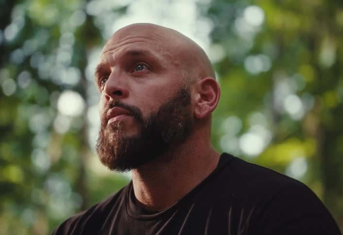 A tough-looking bald man with a beard stands thoughtfully in a sun-dappled forest
