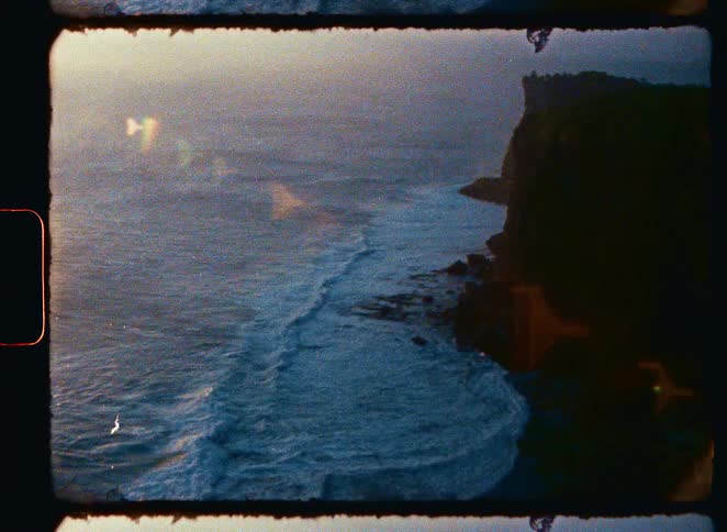 Vintage 8mm film captures ocean waves rolling onto a rocky shore during a hazy sunset
