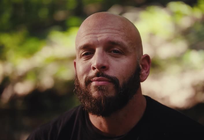 Close-up portrait of a thoughtful bald man with a beard who looks directly into the camera in a forest setting