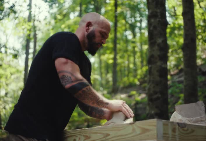 A tattooed, muscular man kneads a large block of clay on a wooden table in a sunlit forest
