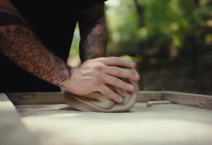Close-up of a tattooed man's hands kneading and preparing clay for pottery in a tranquil forest workshop