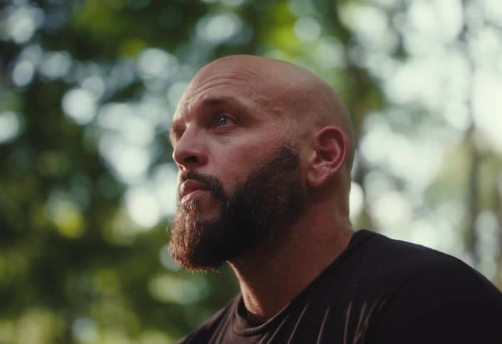 A serious, bald man with a full beard takes a reflective pause in the woods