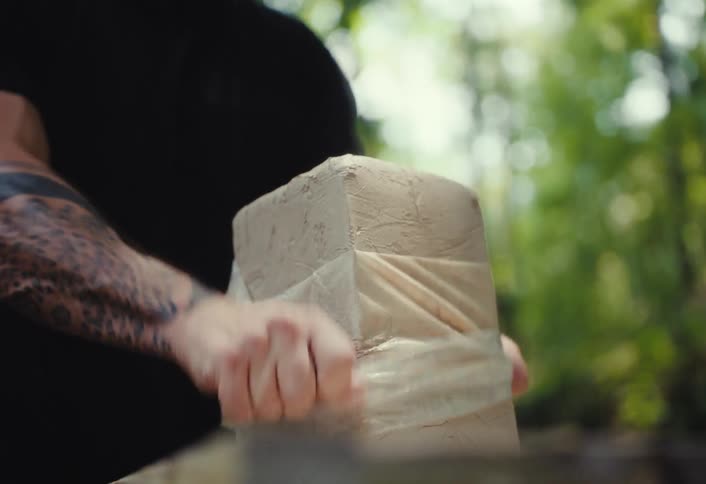 A tattooed male artist unwraps a fresh block of modeling clay to begin sculpting in a serene forest setting