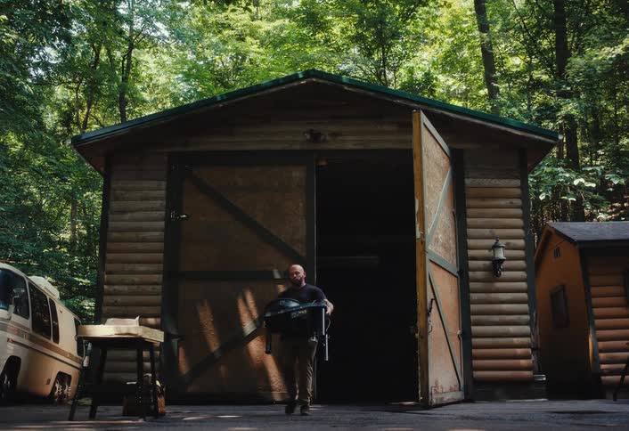 A man in slow motion carries a portable grill out of a rustic wooden garage in the woods