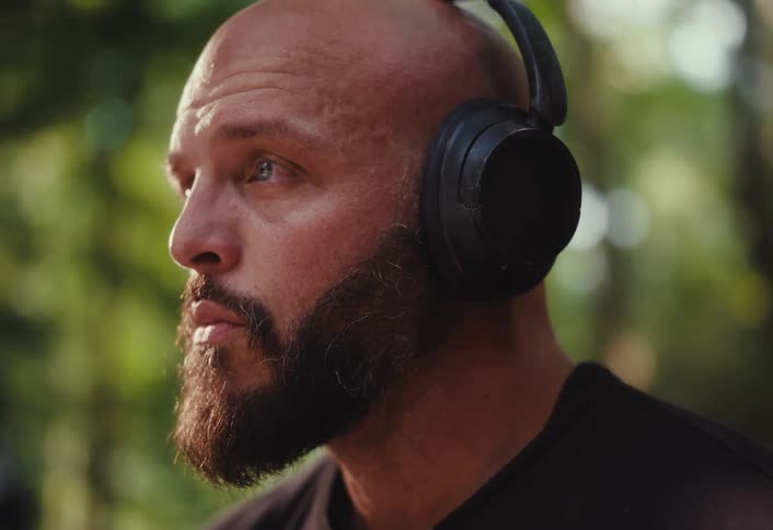 An intense, bald man with a beard wearing headphones listens carefully while outside in a park