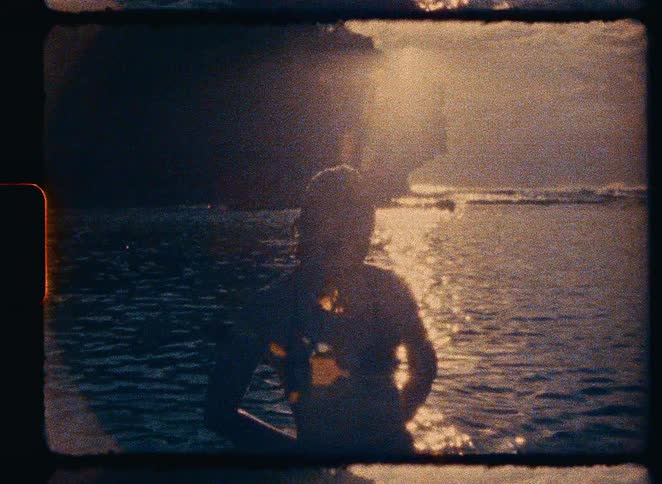 Vintage Super 8 footage captures a woman in a swim cap silhouetted by the golden sunset as she wades in the ocean