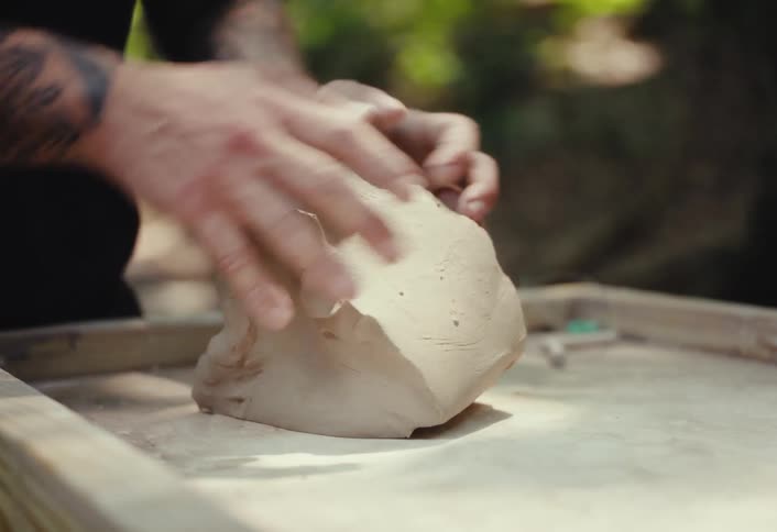 Close-up of a tattooed artist's hands working and kneading a block of fresh clay outdoors in a wooded area