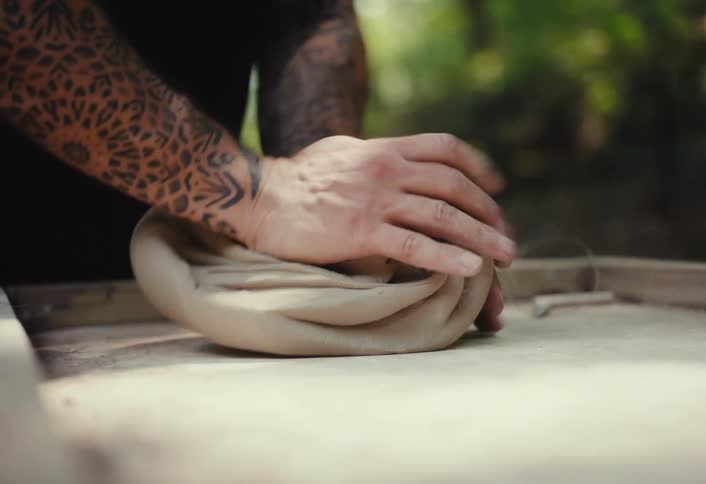 A skilled potter with intricate tattoos on his arms kneads a large piece of clay on a wooden table in his outdoor workshop