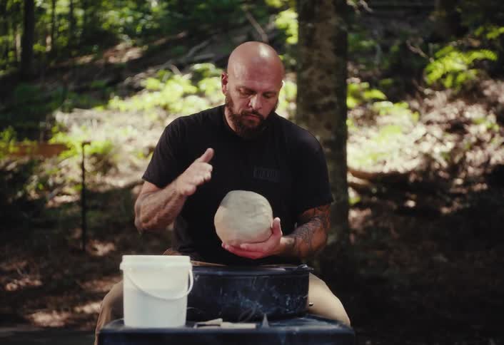 A muscular, tattooed man with a beard centers a lump of clay on a potter's wheel in a serene forest setting