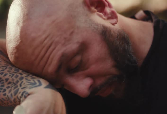 A close-up of an emotionally drained man with a beard resting his weary head on his tattooed arm