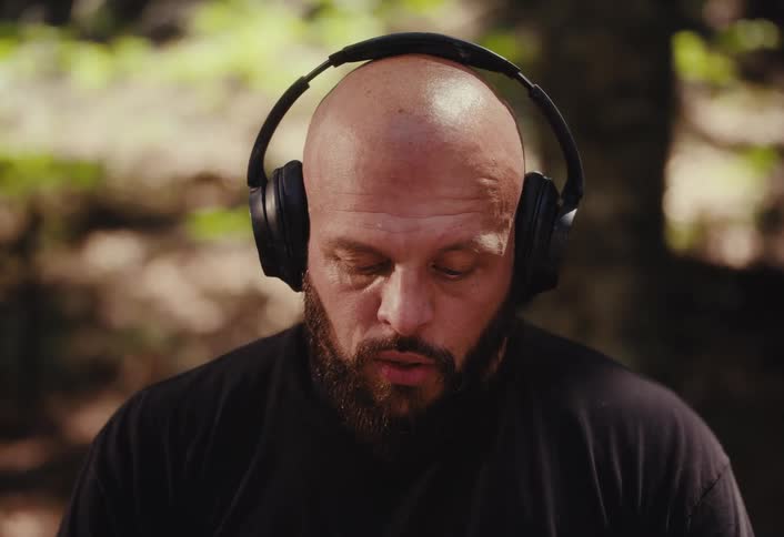 An intense bald man with a beard puts on headphones in a forest and then stares directly into the camera