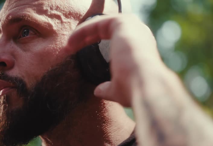 A bald, bearded man wearing headphones takes a deep breath and adjusts them in slow motion during an outdoor workout