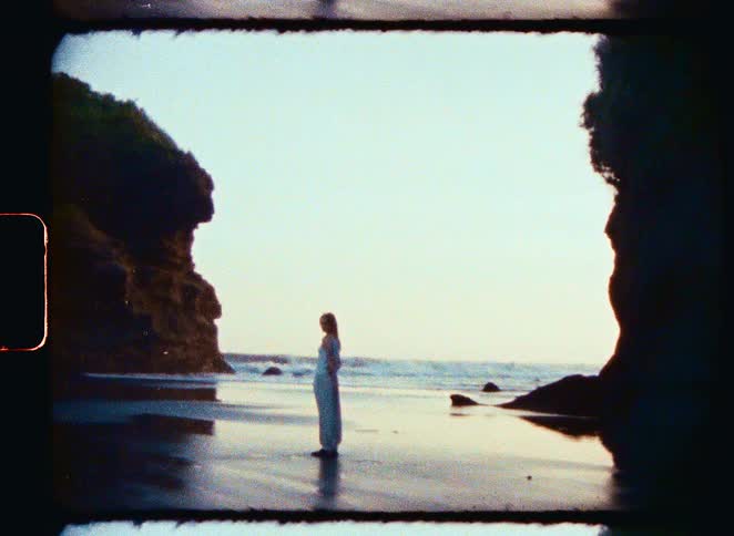 A woman stands silhouetted on a serene beach cove at sunset in vintage 8mm film footage