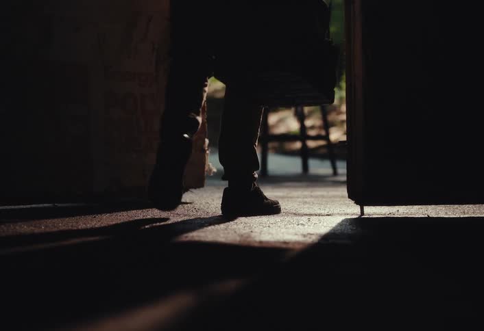 A man's silhouette is revealed as a door opens, and he walks out of a dark garage into the light carrying a tool bag