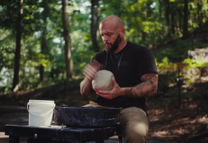 A focused, tattooed man with a beard works with a large lump of clay on his pottery wheel in a peaceful forest setting