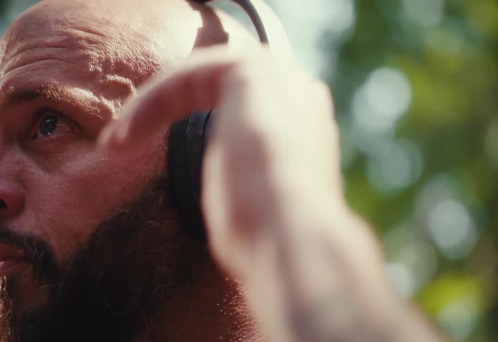 A focused, bald, bearded man listens intently to music on his headphones while outdoors on a sunny day