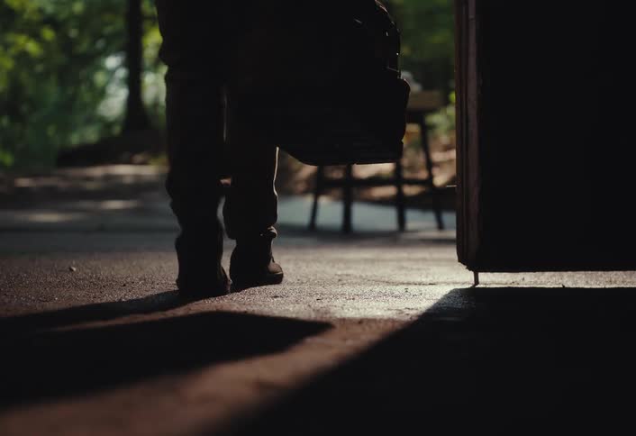A low-angle view follows a silhouetted person's feet as they walk out of a dark room and into a sunlit forest