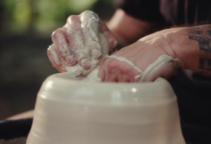 Close up of a potter's hands with tattoos shaping a clay pot on a spinning pottery wheel