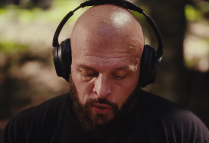 Close-up of a bald, bearded man putting on headphones in a forest and looking up with wide, intense eyes