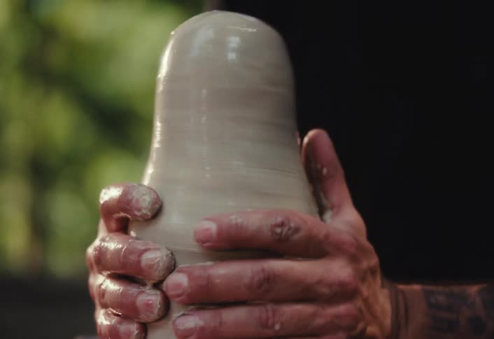 A male potter's hands carefully shape a piece of clay on a spinning pottery wheel in a sunlit workshop