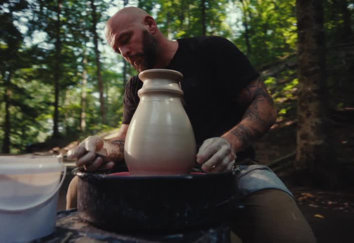 A tattooed artisan carefully shapes a large clay vase on a pottery wheel in a serene, sunlit forest setting