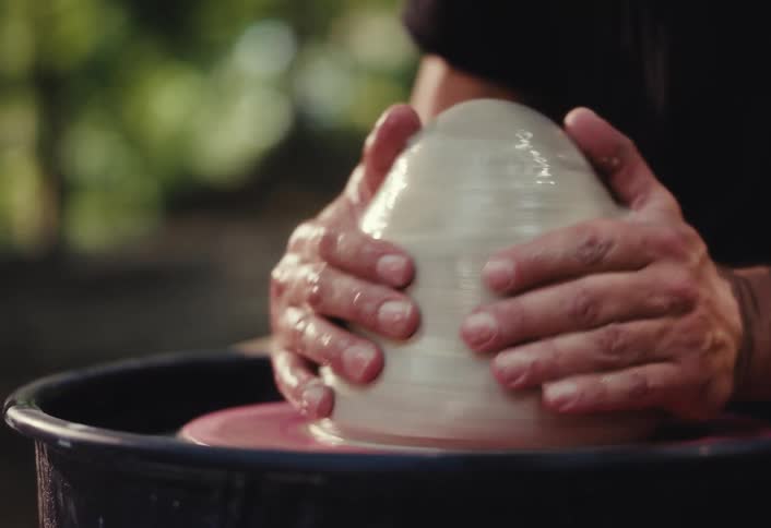 A close-up slow-motion shot shows a potter's strong hands shaping a mound of wet clay on a spinning pottery wheel outdoors