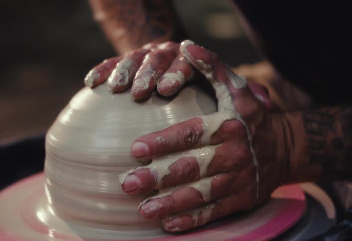 Close up slow motion of a tattooed artisan's hands carefully shaping wet clay on a spinning potter's wheel