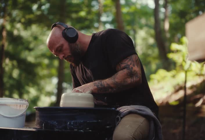 A tattooed man wearing headphones intently shapes clay on a pottery wheel in a tranquil outdoor forest setting