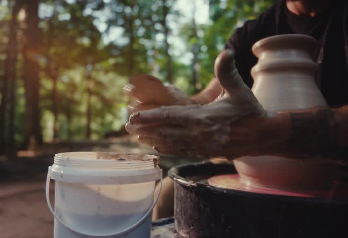 An artisan potter with tattooed arms skillfully crafts a clay pot on a wheel amidst the serene beauty of a forest