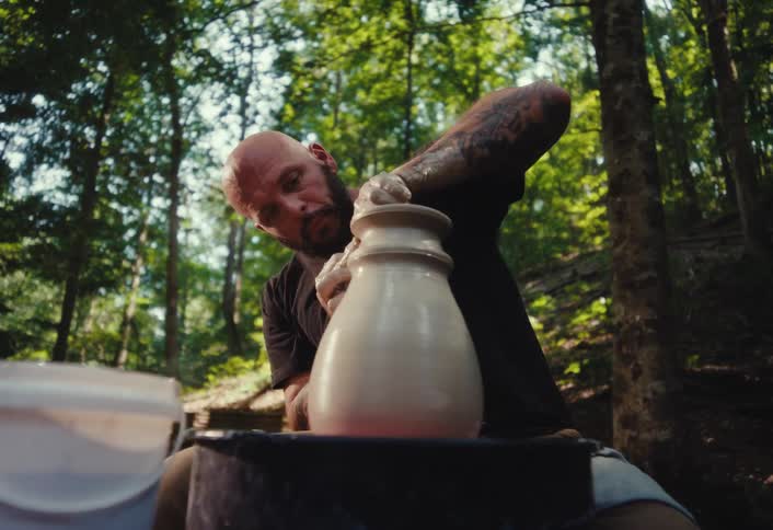 A focused potter with tattoos and a beard shapes a clay vase on his wheel in a serene, sunlit forest