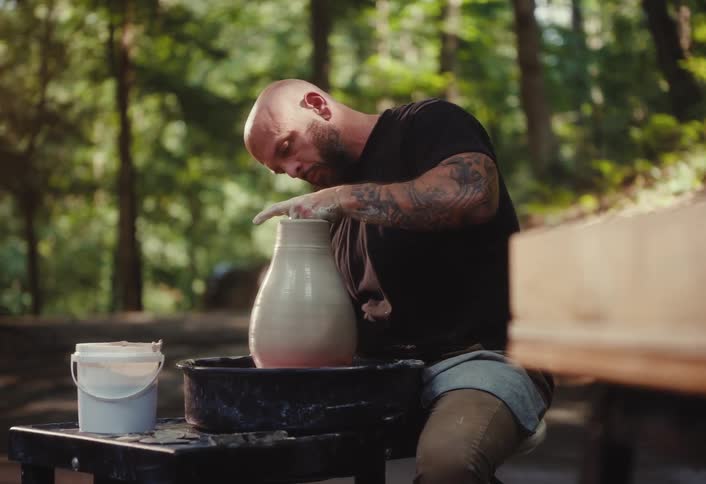 A focused, tattooed male artist skillfully shapes a clay vase on a pottery wheel in a serene forest setting