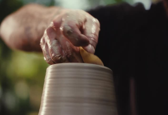 An artisan potter carefully molds and shapes a clay pot on a spinning wheel in a serene outdoor setting