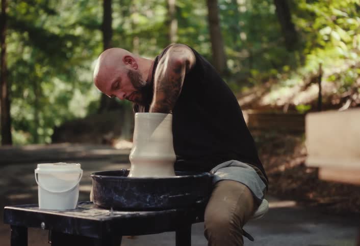 A focused, bearded man with tattoos carefully shapes a large clay vase on a pottery wheel outdoors