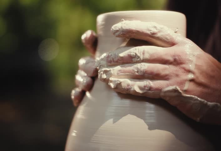 A potter's hands covered in clay meticulously shape a vase on a spinning pottery wheel outdoors