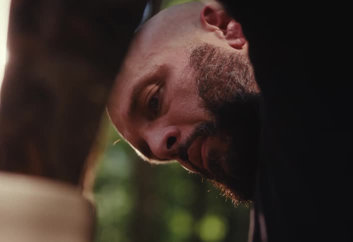 A vertical close-up of a bald, bearded man looking down with intense focus, backlit by the sun