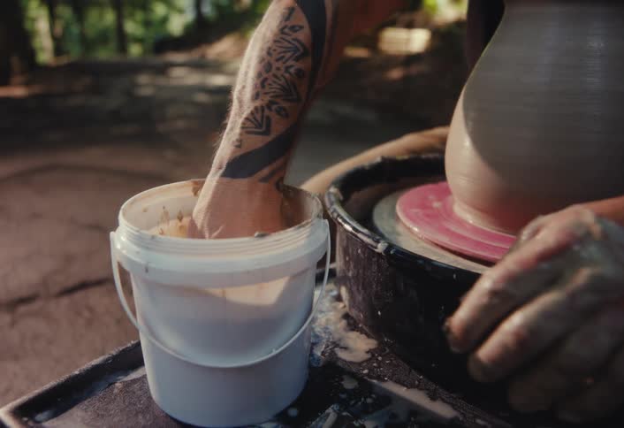Close-up of a potter with tattoos using a sponge and water to shape a clay vase on a pottery wheel outdoors